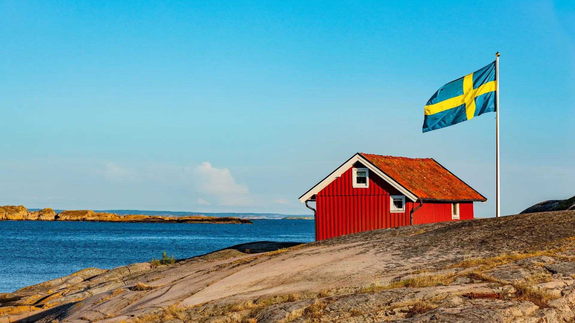 En klassisk röd stuga med svensk flagga vid kusten som representerar Designcements rikstäckande nätverk av certifierade applicerare för microcement i hela Sverige.